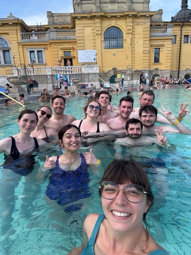Un groupe WeRoad prend un selfie en profitant d'un bain thermal extérieur devant un grand bâtiment historique jaune.