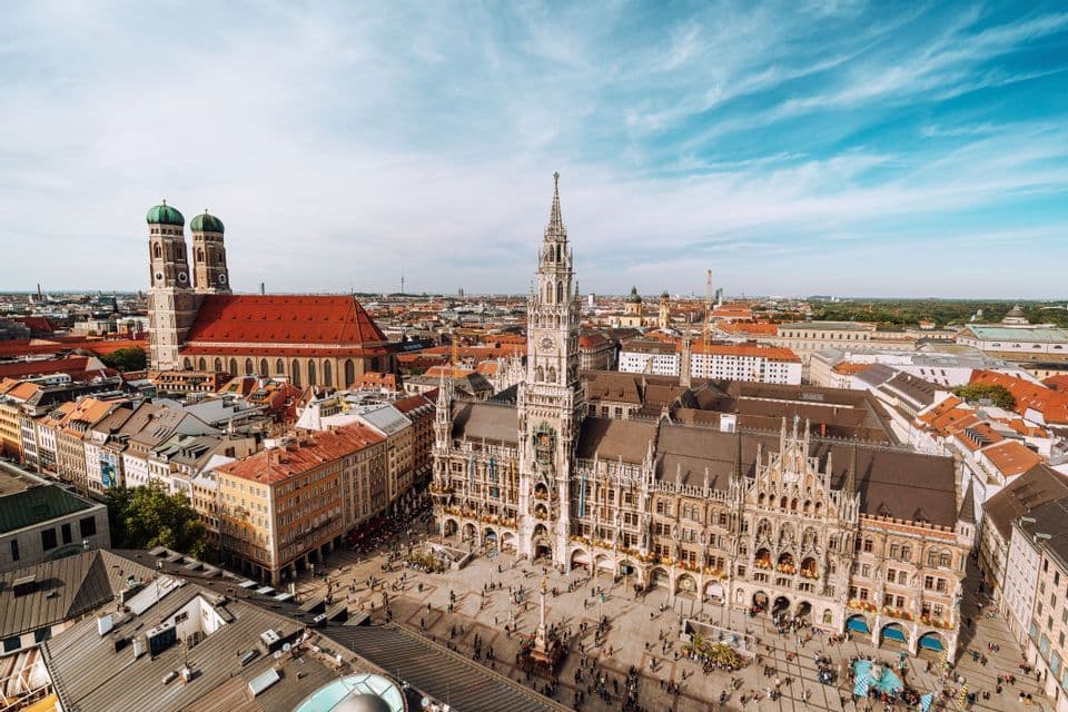 A high-angle view of a historic European city square, featuring an ornate town hall and a twin-domed cathedral under a blue sky.
