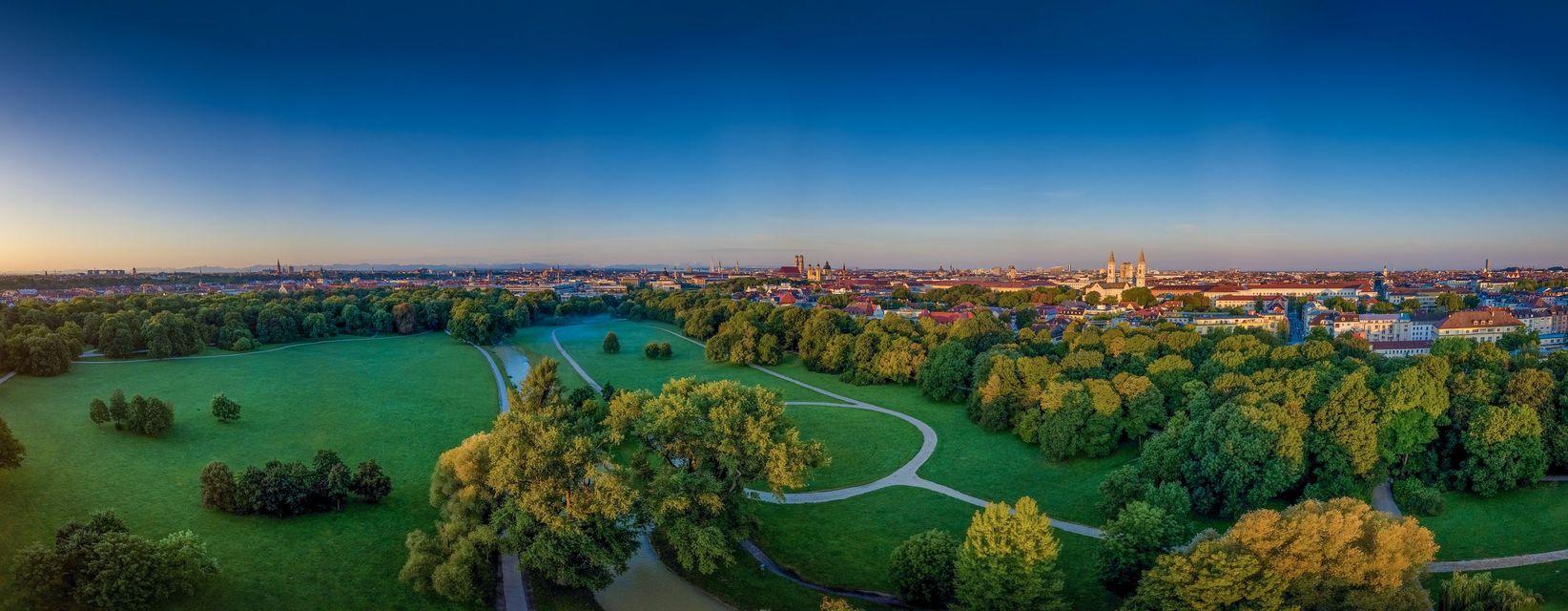 A panoramic aerial view of a large green park with trees and paths in front of a sprawling city skyline under a clear blue sky.