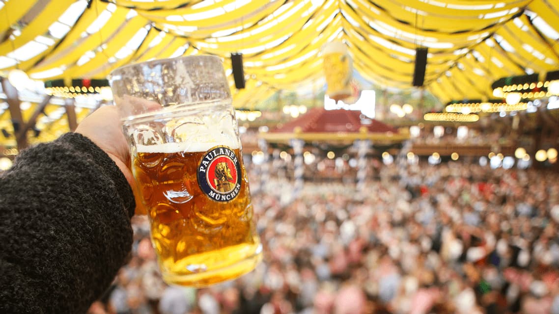 A hand holds up a large glass stein of beer inside a crowded festival tent with a yellow striped ceiling.