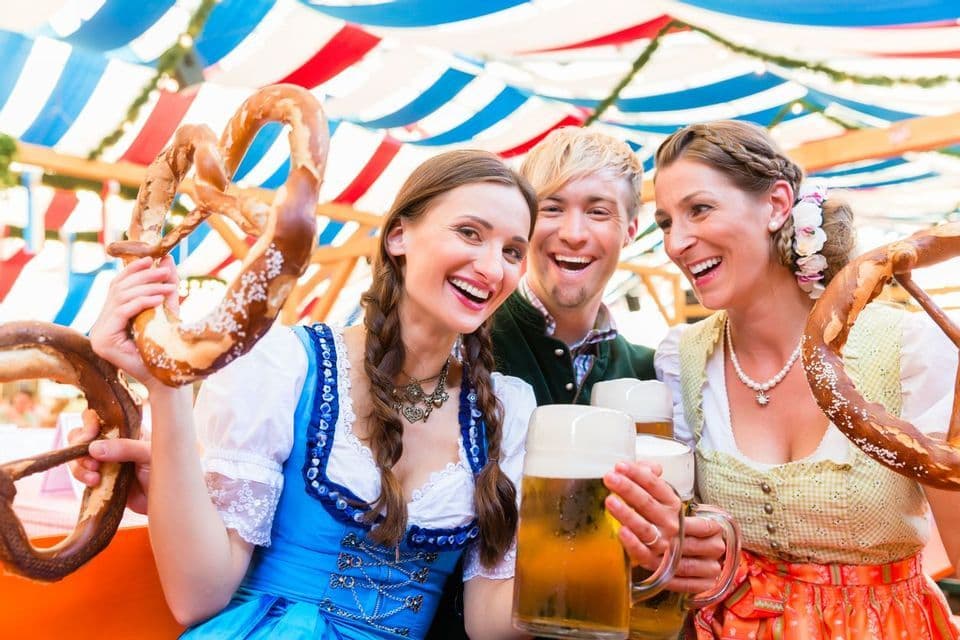 A WeRoad group trip of three friends in traditional Bavarian outfits, laughing while holding large pretzels and beer mugs.