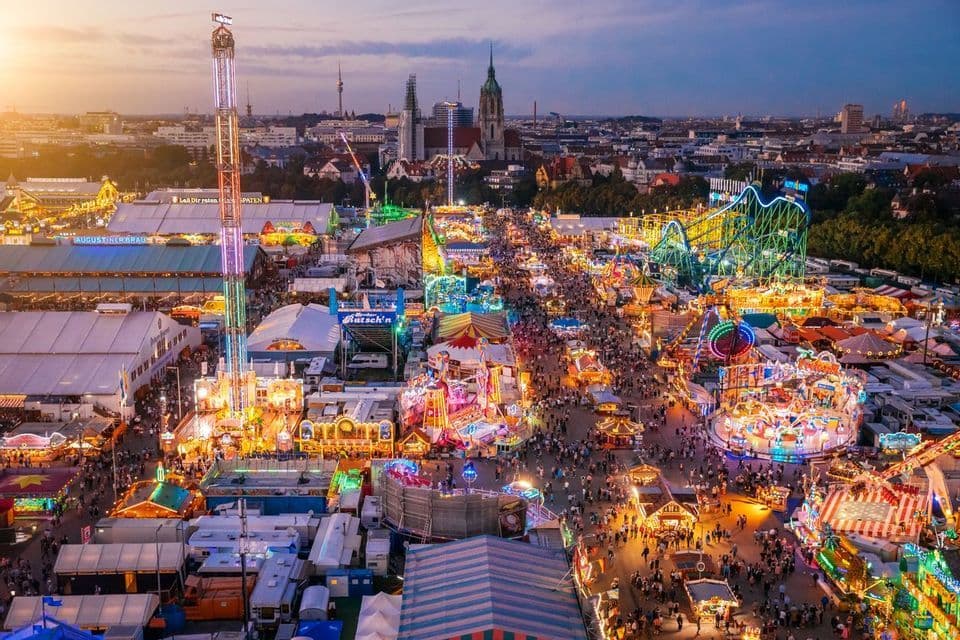 An aerial view of a crowded fairground with glowing rides and attractions at dusk, with a city skyline in the background.