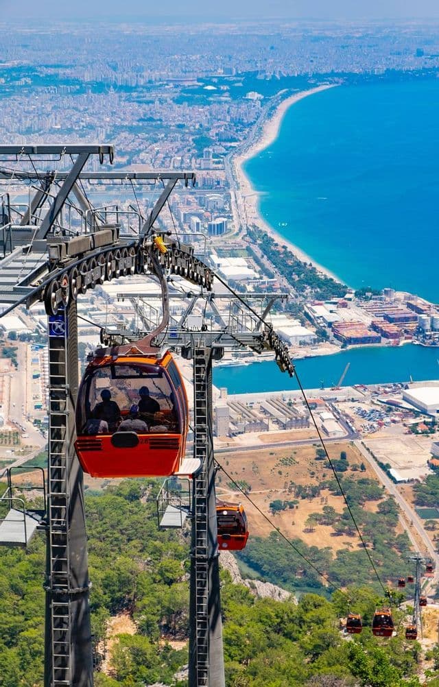 An aerial view from an orange cable car traveling high above green hills, with a sprawling coastal city and blue sea in the background.