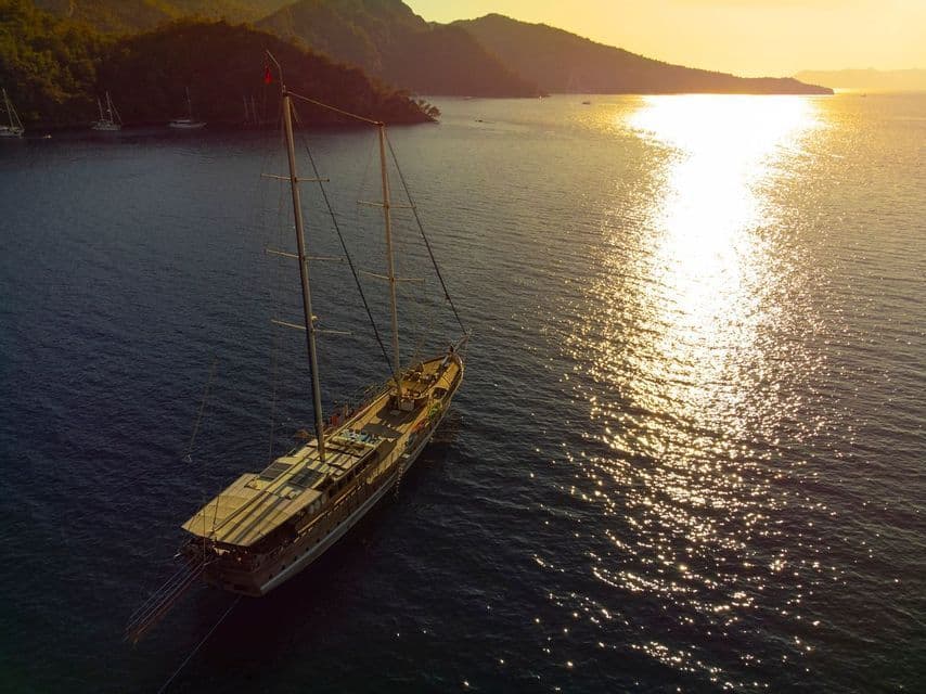 An aerial view of a large wooden sailboat anchored in a bay at sunset, with golden light reflecting on the water.
