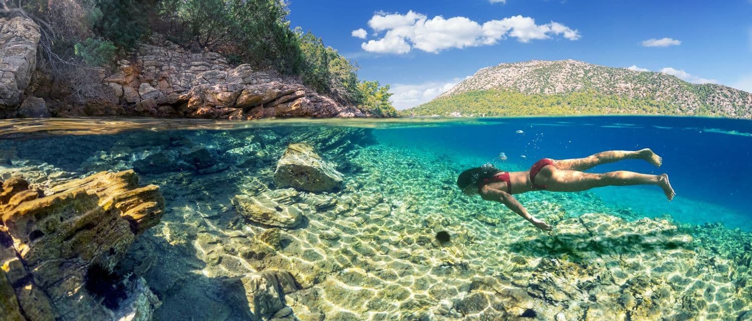 A split-shot of a person in a red bikini swimming underwater along a rocky shoreline under a blue sky.