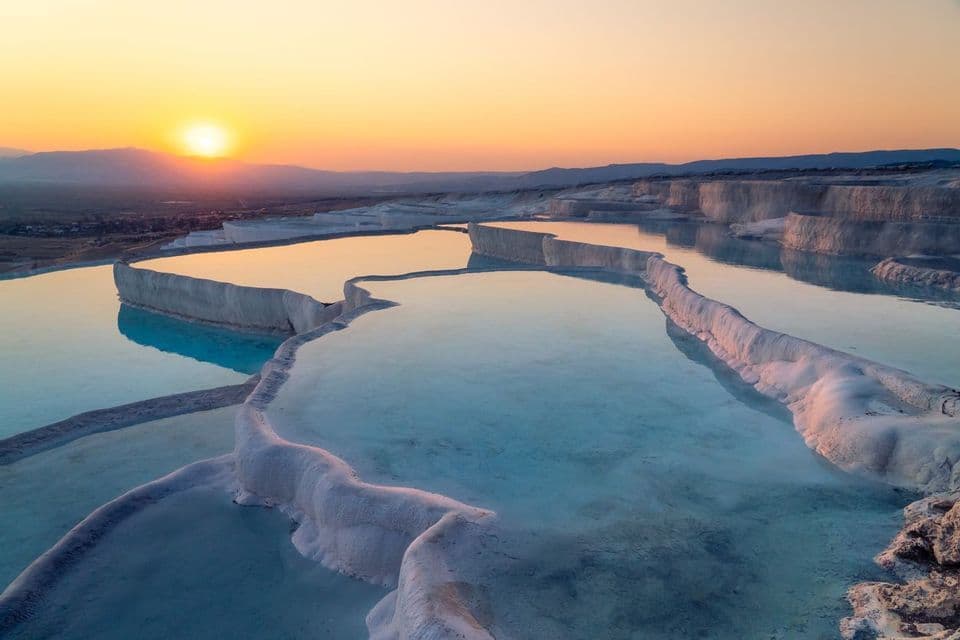 White travertine terraces filled with calm, clear water reflect a golden sunset over distant mountains.