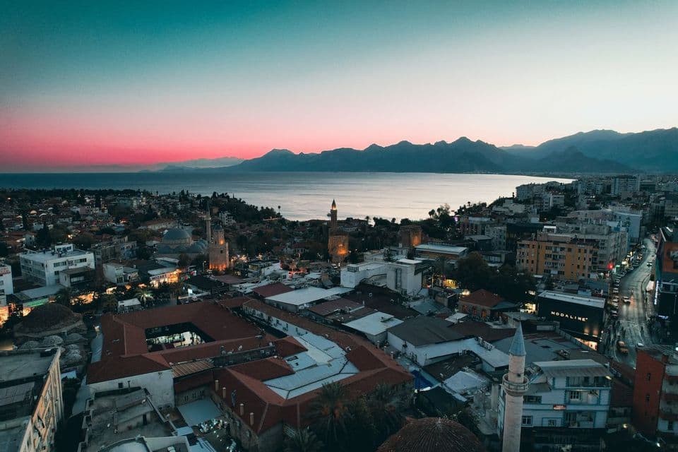 An aerial view of a seaside city with minarets and mountains under a pink and teal sky at dusk.