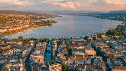 Una vista aerea di una città con un canale centrale che porta a un vasto lago con barche a vela al tramonto.