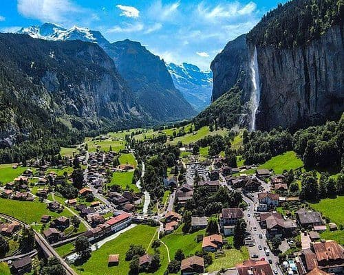 Una vista aerea di un villaggio in una valle verde e lussureggiante, affiancato da ripide montagne con cime innevate e un'alta cascata.