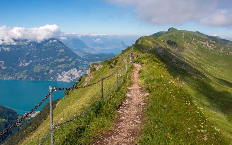 A person hikes along a narrow dirt path on a grassy mountain ridge, with a chain-link fence on the side overlooking a turquoise lake.