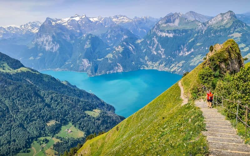 A hiker with a red backpack walks down a stone path on a grassy mountainside overlooking a turquoise lake and distant snow-capped peaks.