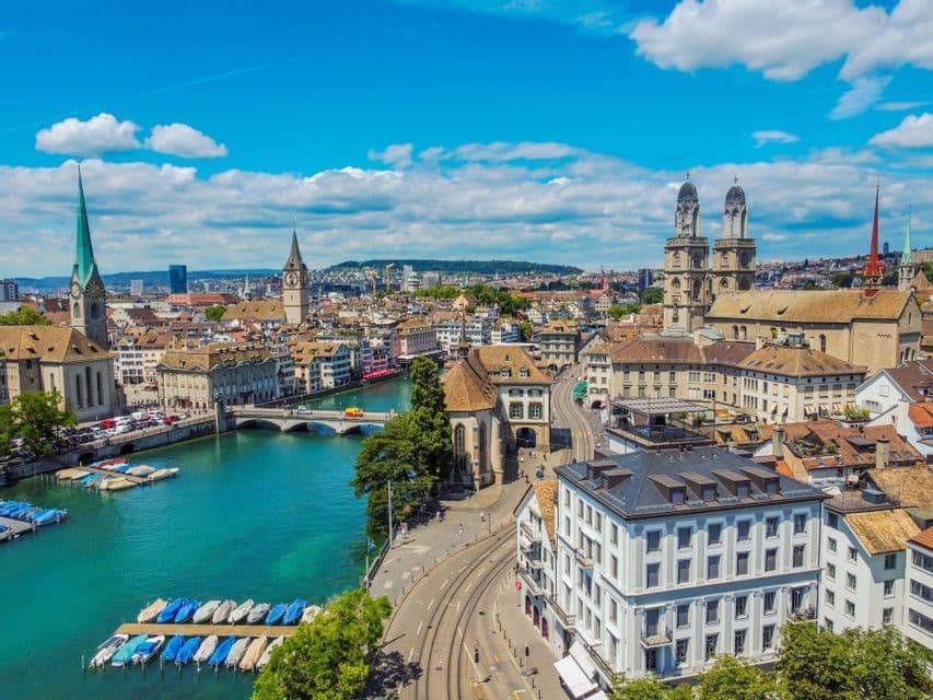 An aerial view of a historic European city with a turquoise river flowing through it, featuring church spires and bridges under a blue sky.