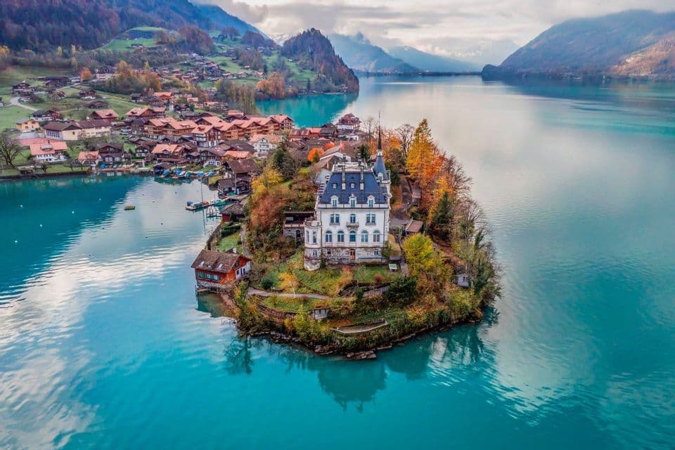 An aerial view of a white castle on a small, tree-covered peninsula jutting into a turquoise lake, with a village and mountains in the background.