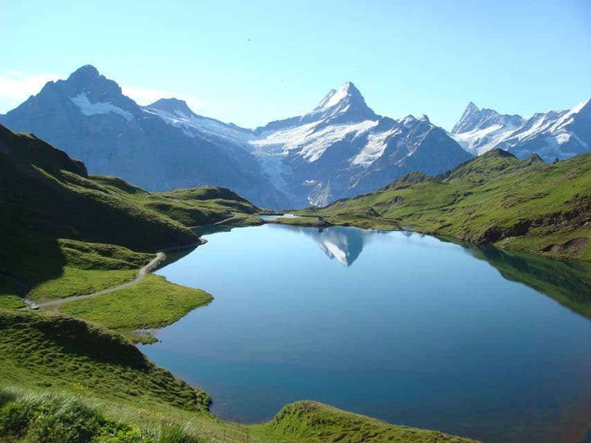 A calm alpine lake reflects snow-capped mountain peaks under a clear blue sky, surrounded by green, rolling hills.
