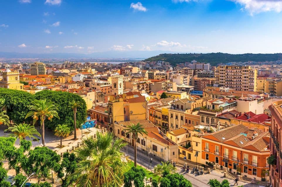 Una vista aérea de una ciudad costera soleada con edificios de colores cálidos, palmeras y colinas distantes bajo un cielo azul.