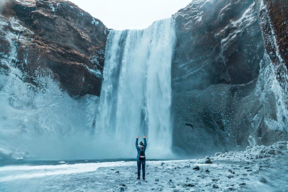 Une personne, les bras levés, fait face à une immense cascade dévalant des falaises sur un rivage glacé.