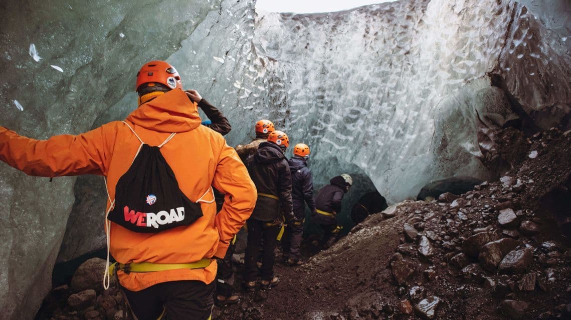 A WeRoad group trip wearing helmets and warm jackets walks in a single file line through a narrow ice cave.