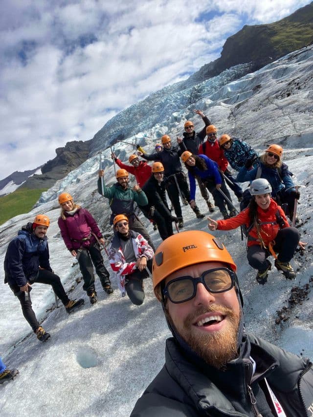 Un homme prend un selfie avec son groupe de voyage WeRoad, tous vêtus de casques orange, posant sur un vaste glacier lors d'un trek en montagne.