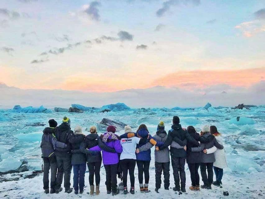 Eine WeRoad-Gruppenreise steht Arm in Arm an einem verschneiten Ufer und blickt auf eine Gletscherlagune mit blauen Eisbergen unter einem Pastellhimmel.