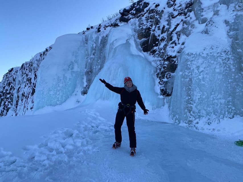 Una donna con casco, imbracatura e ramponi sorride in un paesaggio innevato, di fronte a una grande cascata ghiacciata.