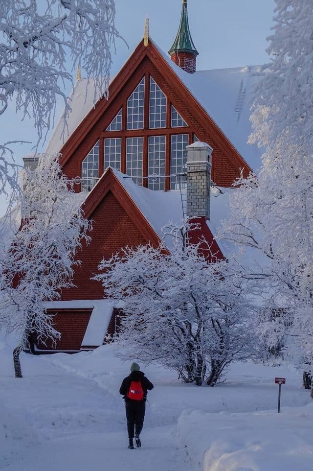 Eine Person mit rotem Rucksack wandert auf einem verschneiten Pfad zu einer großen, roten Holzkirche, umgeben von schneebedeckten Bäumen.