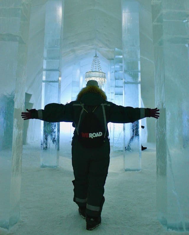 Una persona di spalle con uno zaino WeRoad cammina attraverso un corridoio blu luminoso fatto di pilastri di ghiaccio con le braccia aperte.