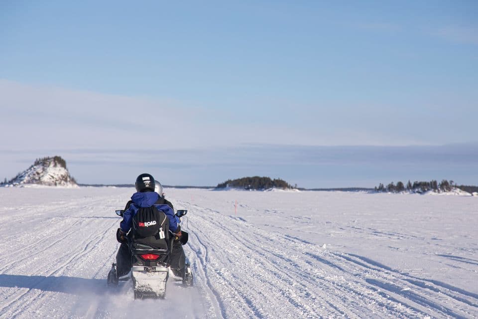 Rückansicht von zwei Personen einer WeRoad Gruppenreise, die mit einem Schneemobil durch eine weite, verschneite Landschaft unter einem klaren blauen Himmel fahren.