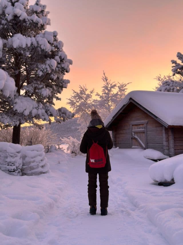 Una persona di spalle con uno zaino WeRoad rosso si trova su un sentiero innevato accanto a una baita di legno al tramonto.