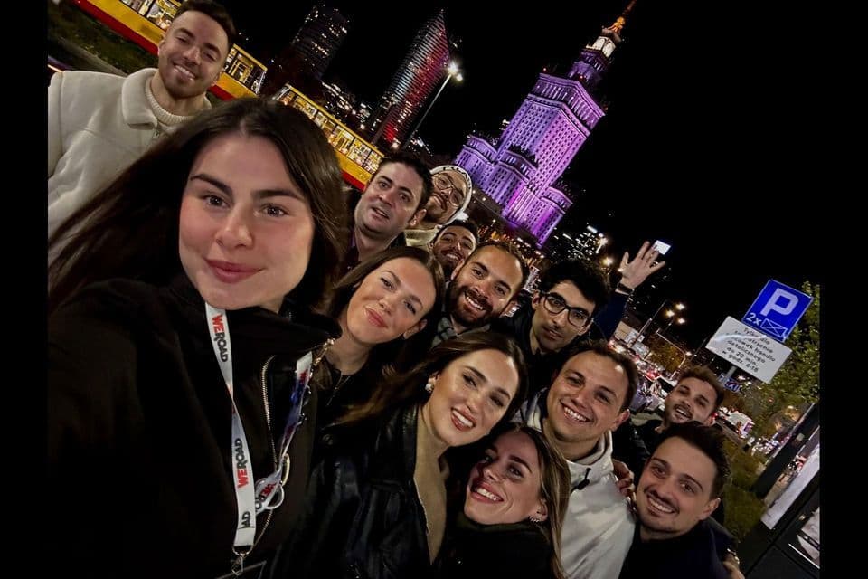 Un viaje en grupo de WeRoad sonríe para una selfie de noche en una ciudad, con un gran edificio iluminado de púrpura al fondo.