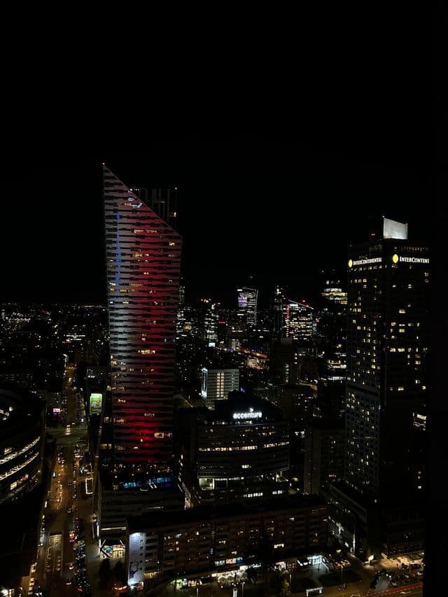 Un extenso horizonte urbano de noche, con innumerables luces de rascacielos y calles abajo. Una torre moderna con techo inclinado está iluminada en rojo.