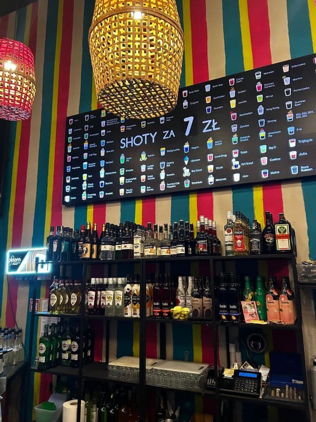El interior de un bar con estantes llenos de botellas de licor debajo de un menú digital que anuncia chupitos frente a una pared de rayas coloridas.