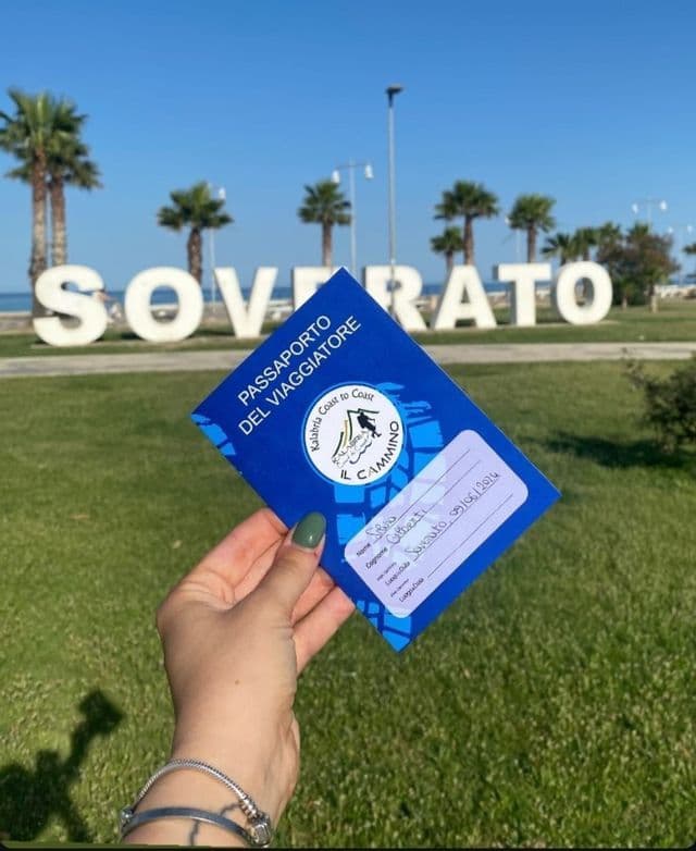 Una mano tiene un passaporto blu 'Kalabria Coast to Coast' davanti a una grande insegna bianca 'Soverato' su un'area erbosa con palme.