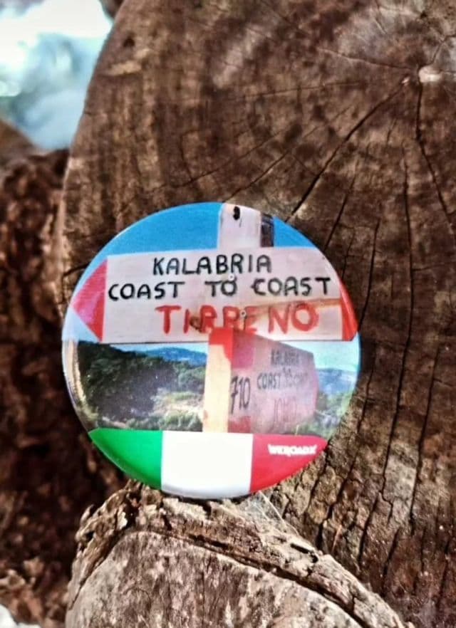 Nahaufnahme eines runden WeRoad-Badges für den 'Kalabria Coast to Coast'-Trail, mit italienischem Flaggendesign, auf einem Holzstamm liegend.