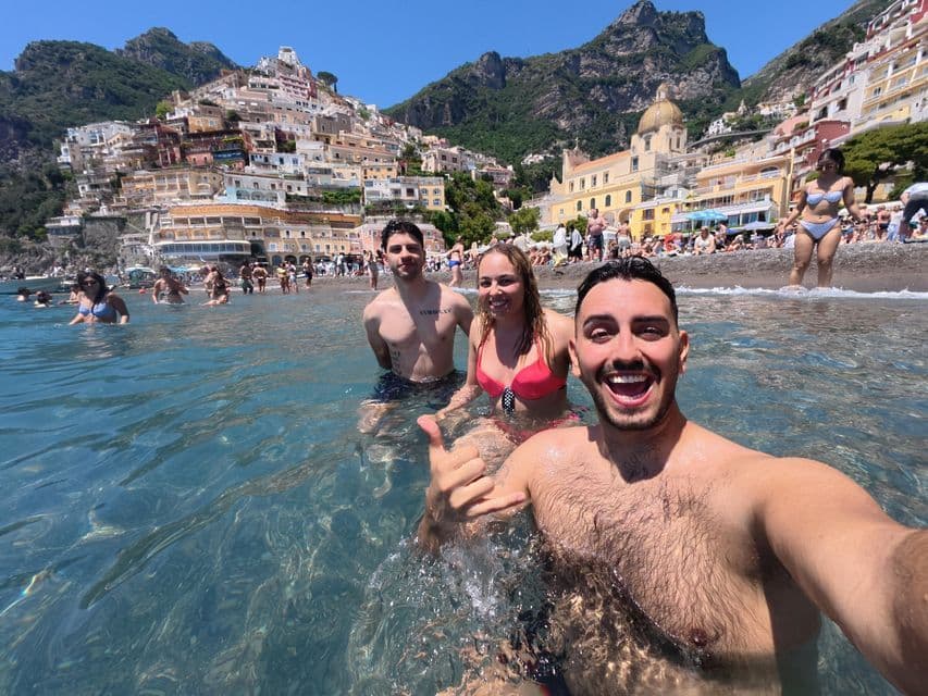 Un uomo di un viaggio di gruppo WeRoad si scatta un selfie con due amici mentre nuota nel mare, con una città collinare affollata sullo sfondo.