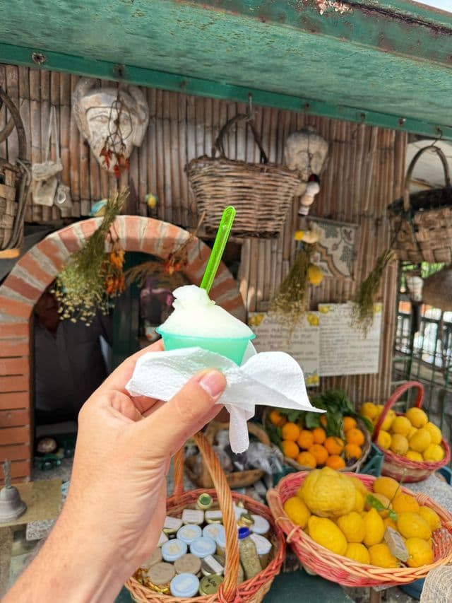 Una mano tiene una coppetta di sorbetto al limone con un cucchiaino verde davanti a una bancarella che vende agrumi freschi.