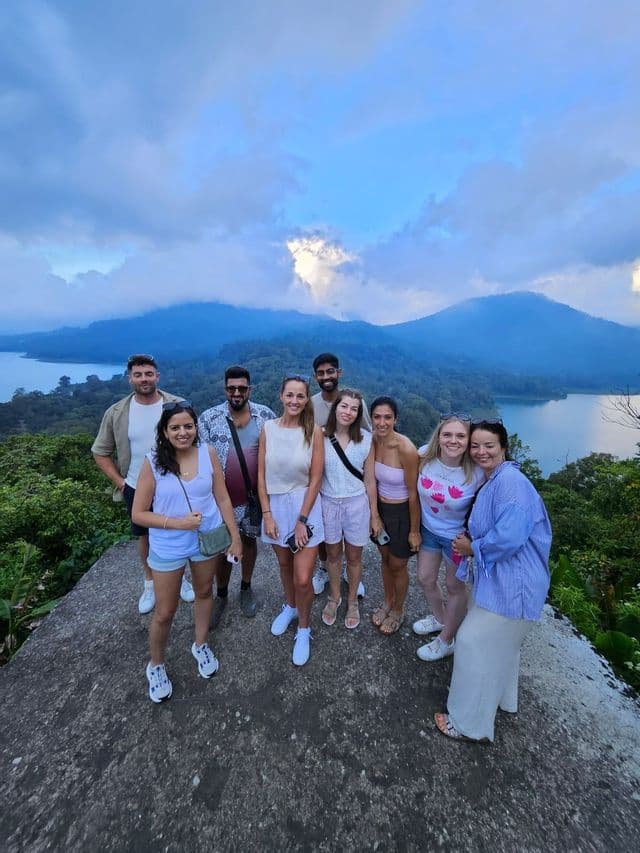 A WeRoad group trip of nine people posing on a viewpoint overlooking a lake and lush mountains under a cloudy sky.