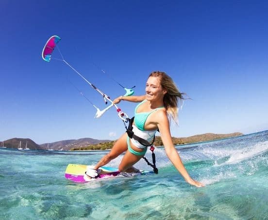 Una mujer sonriente practicando kitesurf en aguas cristalinas de color turquesa, con colinas distantes bajo un cielo azul.