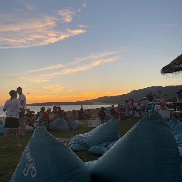 Un viaje en grupo de WeRoad, con personas relajándose en pufs y jugando en el césped junto a la playa durante un atardecer colorido.