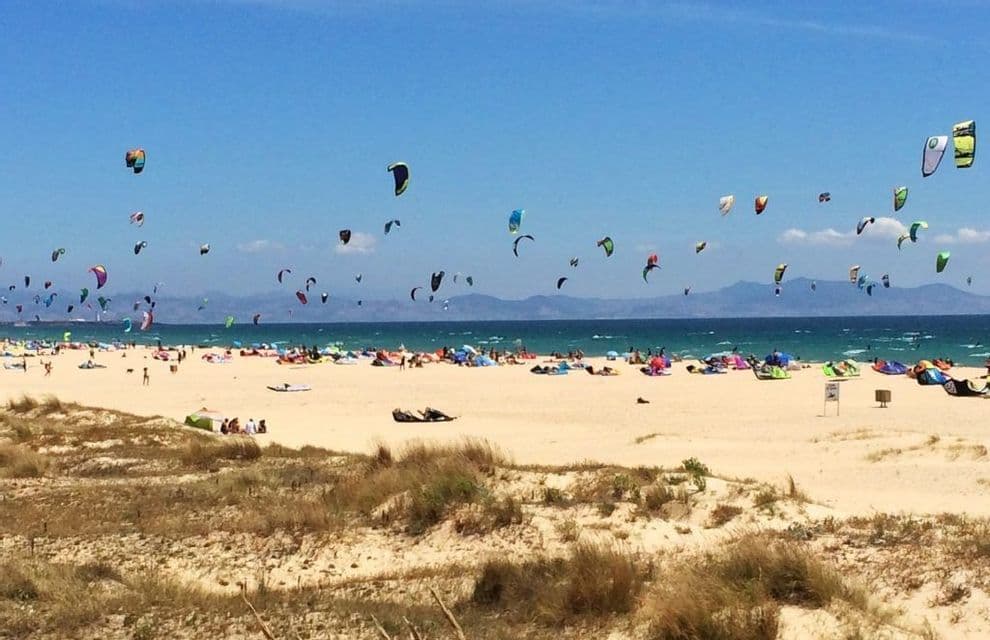 Decenas de cometas coloridas vuelan en el cielo sobre una playa de arena concurrida con gente haciendo kitesurf en el océano.
