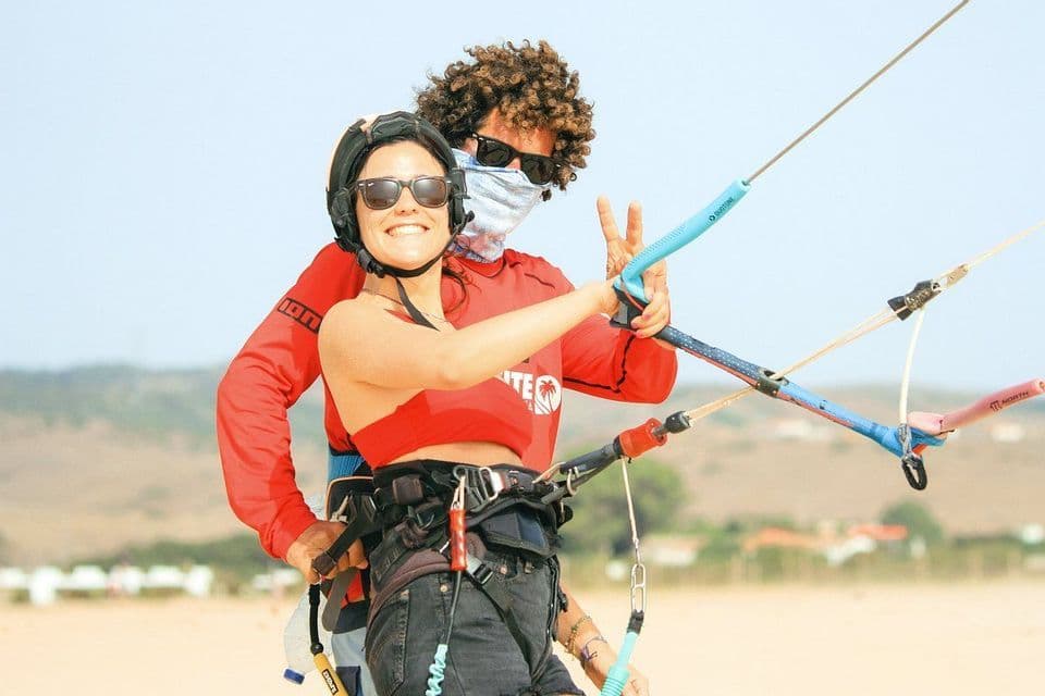 Una mujer con casco sonríe mientras sujeta una barra de control de kitesurf, con un hombre detrás de ella en una playa de arena.