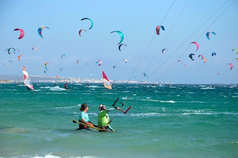 Dos personas en primer plano aprenden a hacer kitesurf mientras muchas otras practican kitesurf y windsurf en un mar turquesa agitado bajo un cielo azul claro.