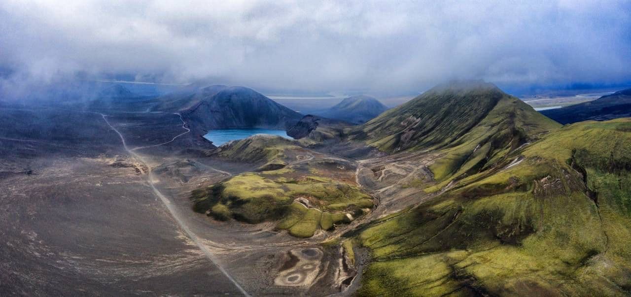 An aerial view of a volcanic landscape with green-covered mountains, a small blue lake, and a winding dirt road under a cloudy sky.