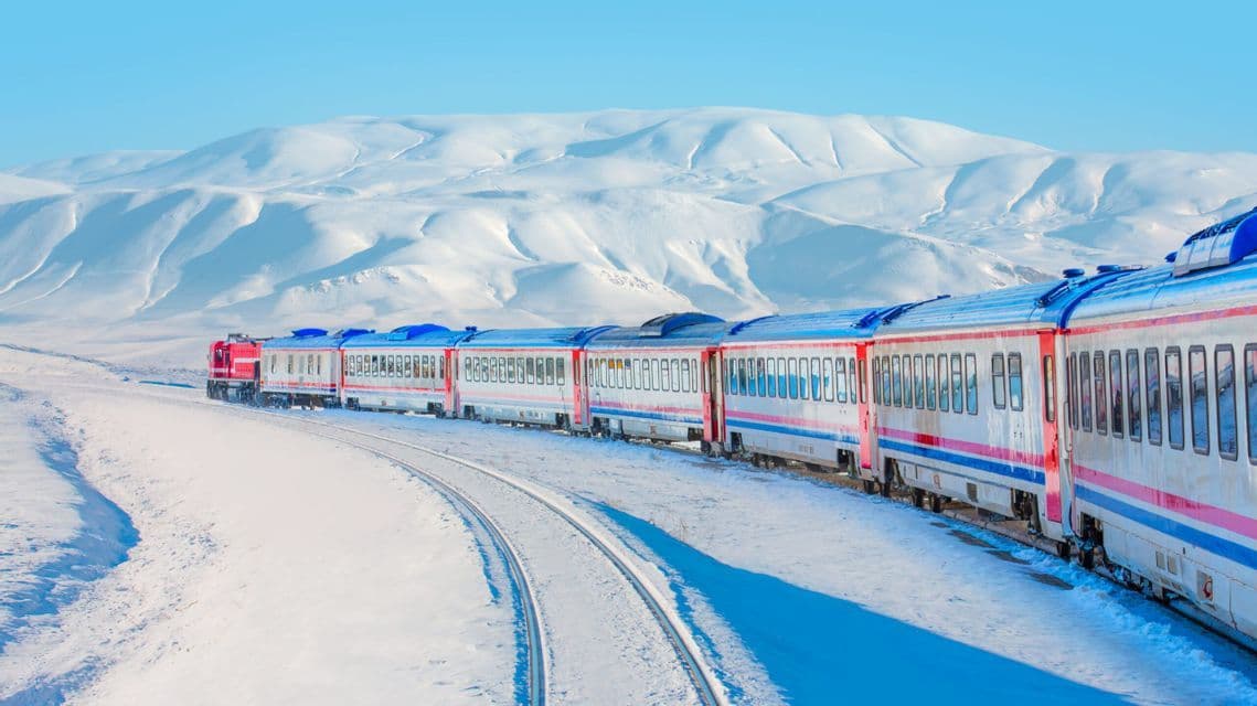 Un treno passeggeri rosso e bianco viaggia su un binario curvo attraverso un paesaggio di montagne innevate sotto un cielo azzurro e limpido.