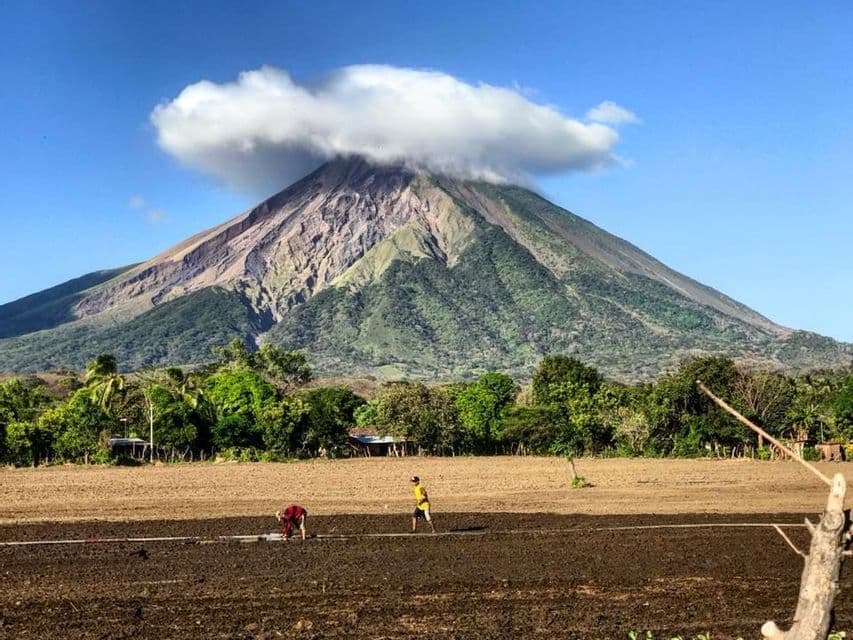 Vue d'un grand volcan au sommet nuageux, avec un champ labouré et des personnes au premier plan.