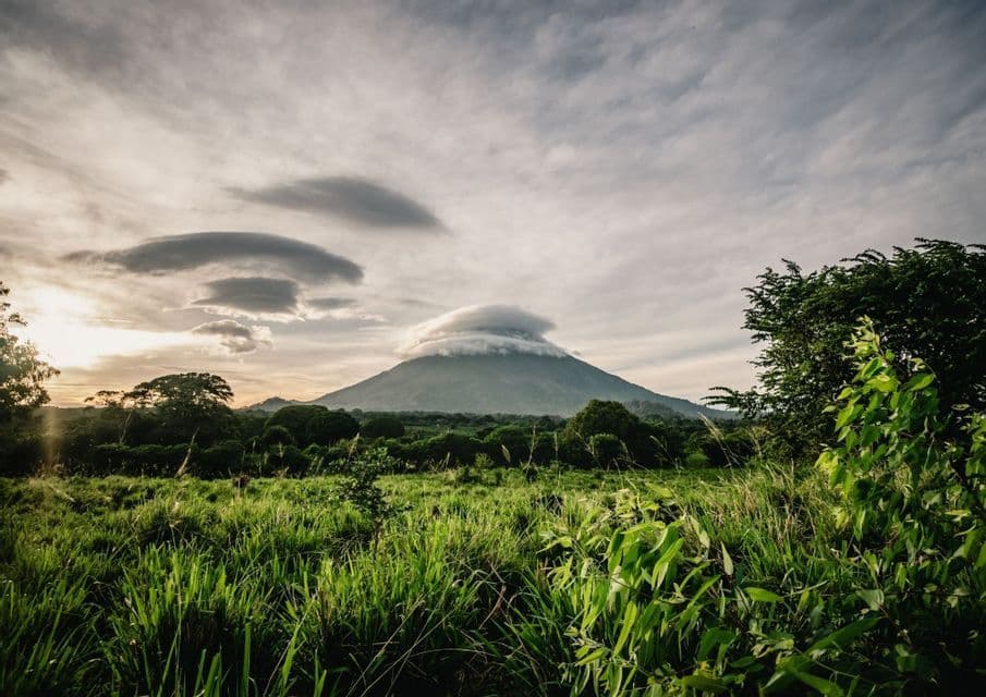Un volcan dont le sommet est couvert par un nuage lenticulaire, vu depuis un champ herbeux sous un ciel nuageux au coucher du soleil.