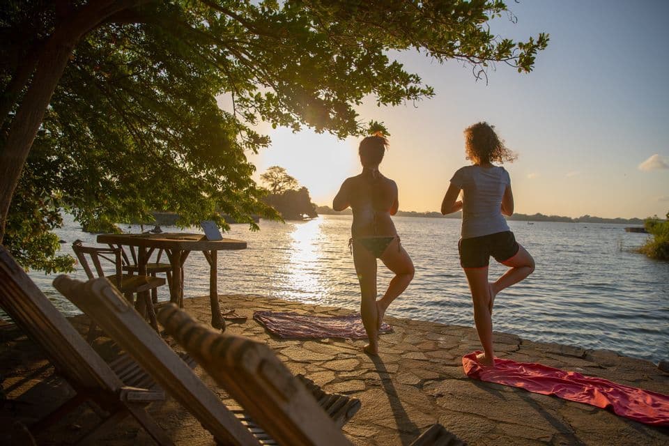 Deux femmes pratiquent le yoga en posture de l'arbre sur un patio en pierre au bord de l'eau, en contre-jour du soleil couchant.
