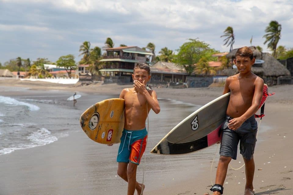 Deux jeunes garçons marchent le long d'une plage de sable, chacun portant une planche de surf sous le bras.