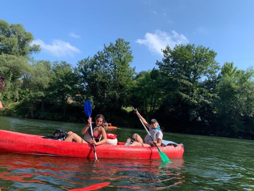 Dos mujeres de un viaje en grupo de WeRoad posando con remos en un kayak rojo en un río, con una orilla arbolada al fondo.