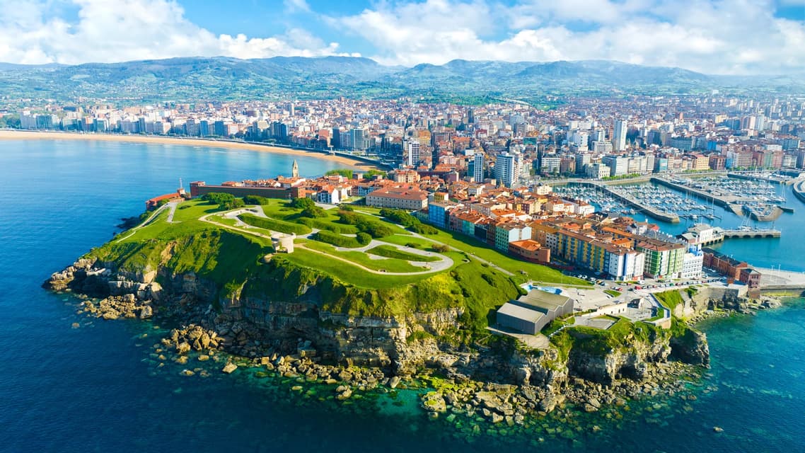 Una vista aérea de una ciudad costera en una península verde, con una gran playa y un puerto deportivo lleno de barcos.