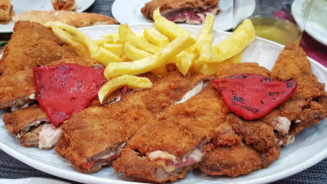 Primer plano de un plato con milanesas de carne empanadas, rellenas de jamón y queso, servidas con patatas fritas y pimientos rojos asados.